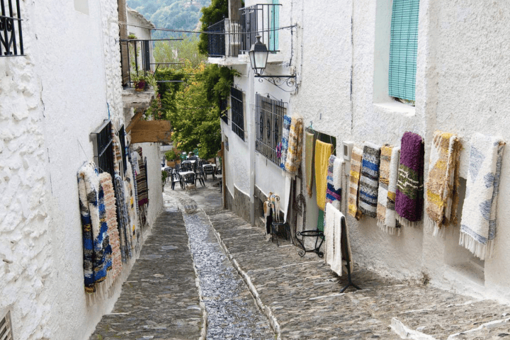 Narrow cobblestone street with colorful rugs hanging on white walls.